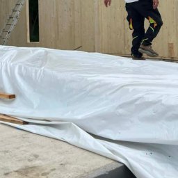 Wasyl Moroc - Montaż domu modułowego w Pliszczynie. Widoczna drewniana konstrukcja ściany z otworem okiennym, zabezpieczona folią. Pracownik wchodzi na konstrukcję.