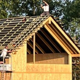 Wasyl Moroc - Montaż drewnianej konstrukcji dachu domu modułowego przez dwóch pracowników w słoneczny dzień. Widoczne elementy więźby dachowej i poszycia.