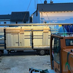 Wasyl Moroc - Transport i montaż prefabrykowanych elementów domu modułowego na naczepie ciężarówki, widoczny dźwig i pracownik w kasku, w tle zabudowa jednorodzinna.