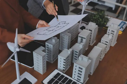 Analiza planu budynku z modelami wieżowców i turbin wiatrowych. Osoby wskazują na projekt, sugerując planowanie przestrzenne lub ocenę energetyczną.