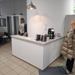 Albert Styl - Wnętrze butiku odzieżowego z białą ladą, manekinem w beżowej kurtce z futrzanym kołnierzem i klientką w przymierzalni w tle.