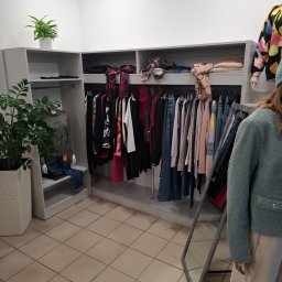 Albert Styl - Wnętrze butiku odzieżowego z szarymi regałami, ubraniami na wieszakach i manekinem ubranym w sweter i dżinsy, uzupełnione roślinami doniczkowymi.