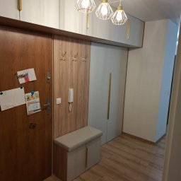 Albert Styl - Przedpokój z zabudową na wymiar: szafa wnękowa z drewnianym panelem z wieszakami, szafka z siedziskiem, białe szafki wiszące z złotymi uchwytami, drewniane drzwi z rysunkami dziecięcymi i lampy...