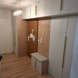 Albert Styl - Przedpokój z zabudową szaf na wymiar w kolorze szarym z długimi złotymi uchwytami, drewnianym panelem z wieszakami i siedziskiem, oświetlony nowoczesną lampą wiszącą.