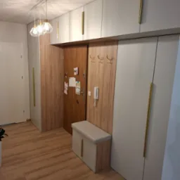 Przedpokój z zabudową szaf na wymiar w kolorze szarym z długimi złotymi uchwytami, drewnianym panelem z wieszakami i siedziskiem, oświetlony nowoczesną lampą wiszącą.