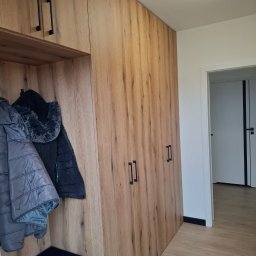 Albert Styl - Wysoka zabudowa w przedpokoju z drewnopodobnymi frontami, czarnymi uchwytami i wnęką z wieszakami na kurtki oraz siedziskiem z czarną czapką.