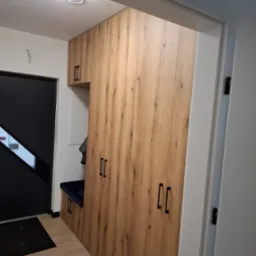 Wysoka szafa z drewnianymi frontami w przedpokoju, z wbudowanym siedziskiem i czarnymi, podłużnymi uchwytami, obok drzwi wejściowych.