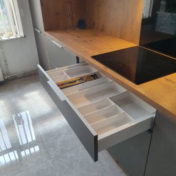 Albert Styl - Otwarta szuflada kuchenna z przegródkami na sztućce i akcesoria, widoczne opakowania folii aluminiowej, blat z płyty imitującej drewno, płyta indukcyjna w tle, szare fronty szafek.