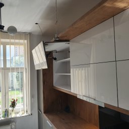 Albert Styl - Nowoczesna kuchnia z białymi, lakierowanymi szafkami wiszącymi i drewnianym blatem, widok na okno z żaluzjami i kwiatami.