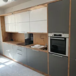 Nowoczesna zabudowa kuchenna z szarymi frontami, białymi szafkami górnymi i blatem imitującym drewno, z wbudowanym piekarnikiem i płytą grzewczą.