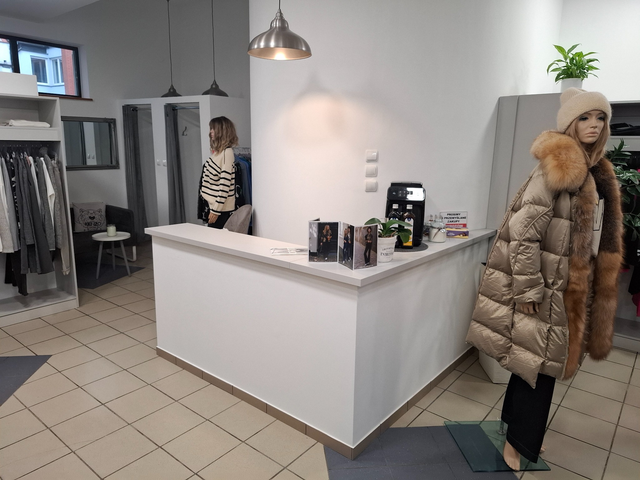 Wnętrze butiku odzieżowego z białą ladą, manekinem w beżowej kurtce z futrzanym kołnierzem i klientką w przymierzalni w tle.