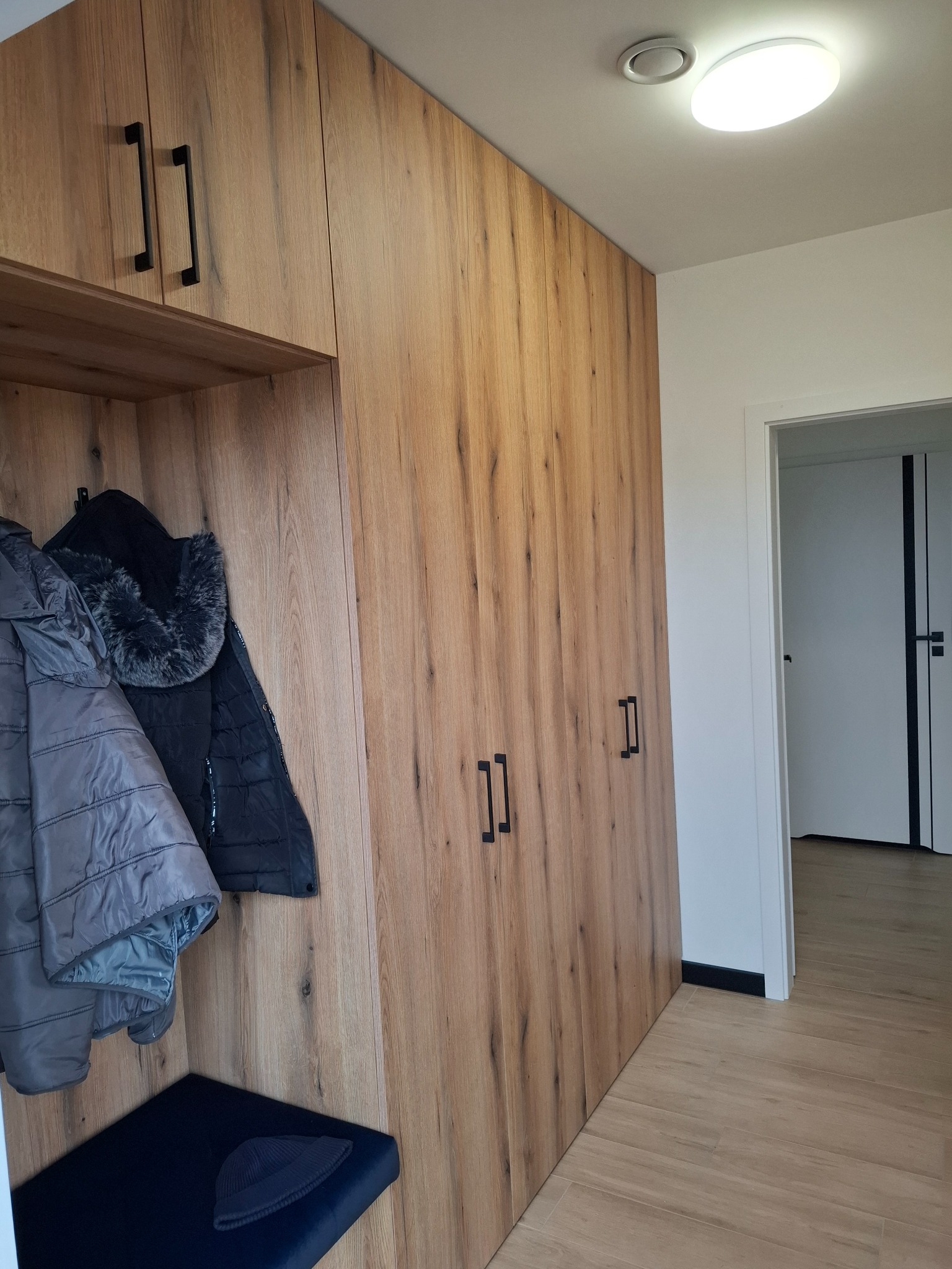 Wysoka zabudowa w przedpokoju z drewnopodobnymi frontami, czarnymi uchwytami i wnęką z wieszakami na kurtki oraz siedziskiem z czarną czapką.