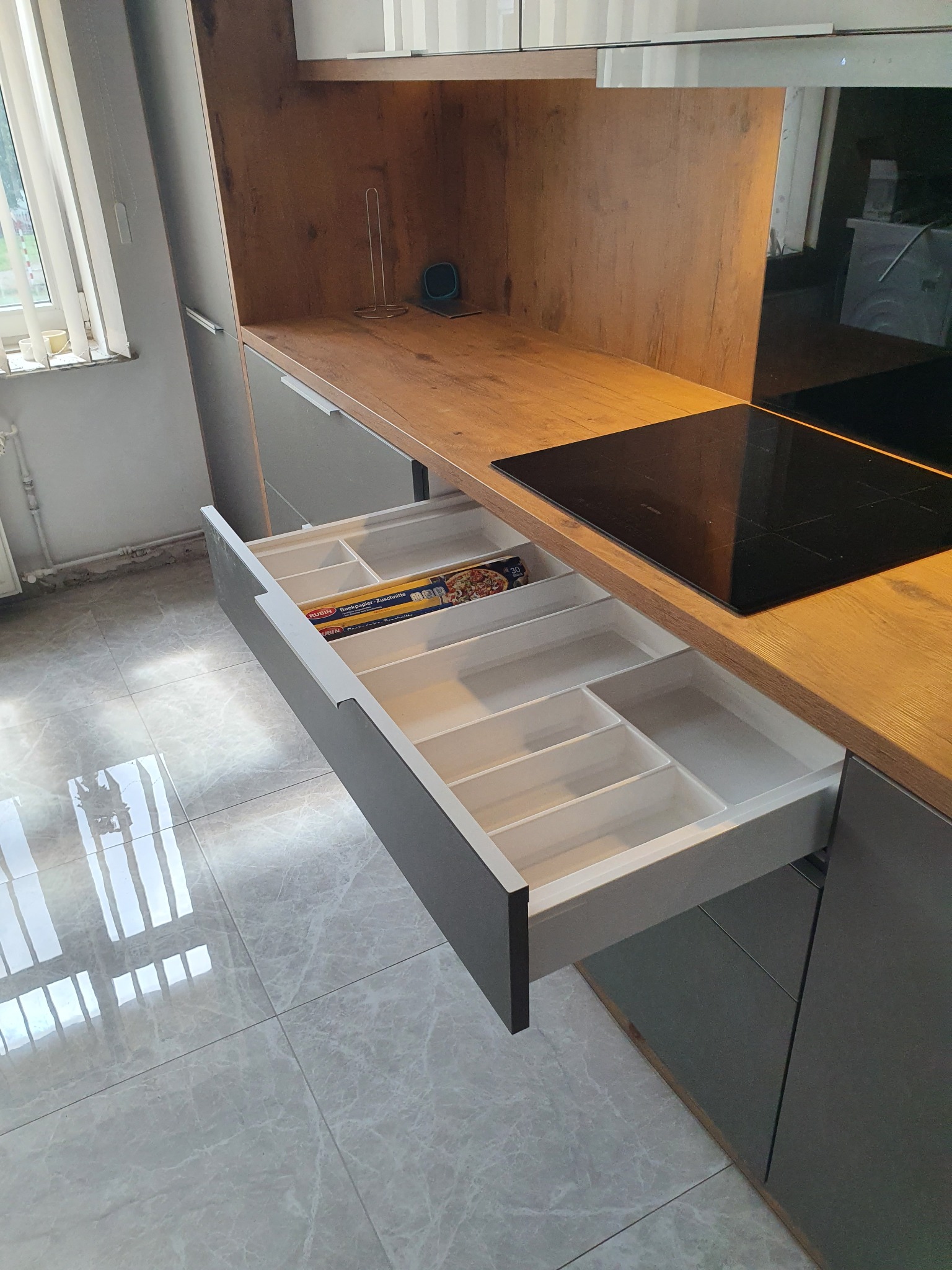 Otwarta szuflada kuchenna z przegródkami na sztućce i akcesoria, widoczne opakowania folii aluminiowej, blat z płyty imitującej drewno, płyta indukcyjna w tle, szare fronty szafek.