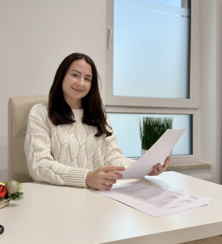 Uśmiechnięta kobieta w białym swetrze siedzi za biurkiem, trzymając dokumenty. W tle okno z rośliną i minimalistyczny wystrój wnętrza. Na biurku świąteczna dekoracja.