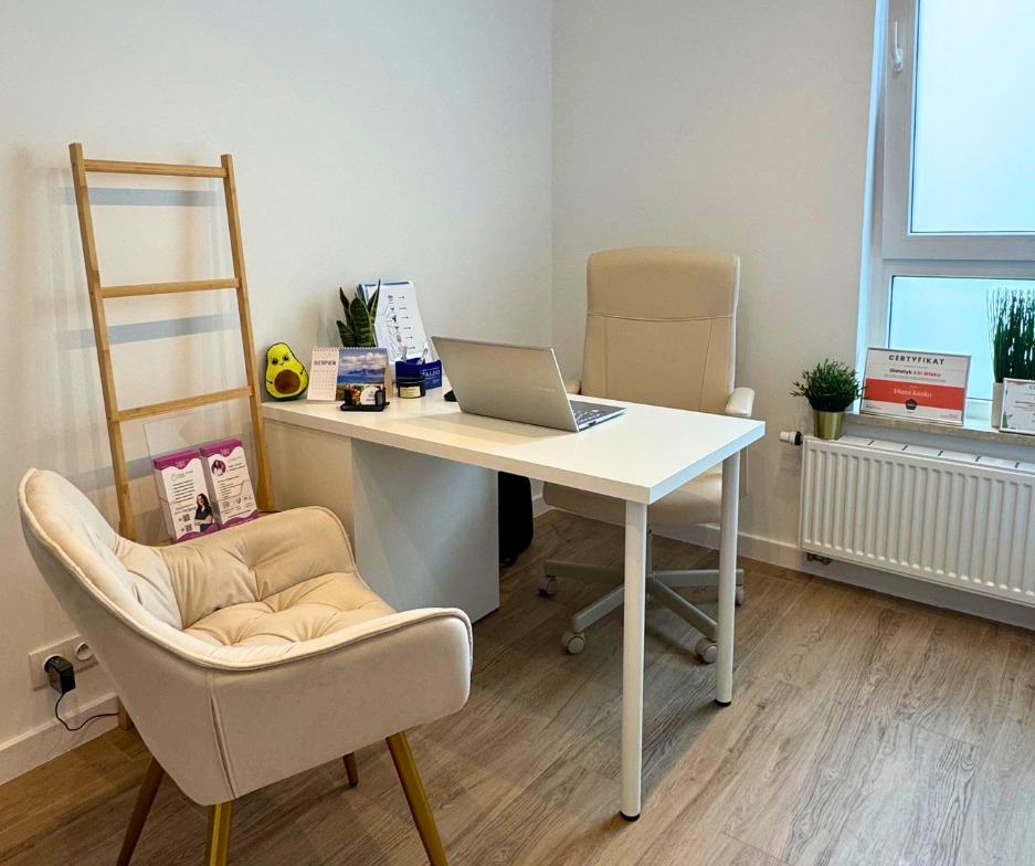 Jasne biuro dietetyka z biurkiem, krzesłami, laptopem i dekoracjami. Na parapecie certyfikat dietetyka. Minimalistyczny wystrój, drewniana podłoga.