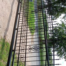 Kamil Jaskólski - Czarna, metalowa brama z dekoracyjnymi elementami w kształcie wolut i rombów, zamontowana na ceglanym murku, widok z perspektywy ulicy.