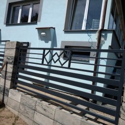 Kamil Jaskólski - Nowoczesne, poziome ogrodzenie metalowe w kolorze grafitowym, z dekoracyjnymi elementami romboidalnymi, zamontowane na betonowej podmurówce przy jasnoniebieskim domu.