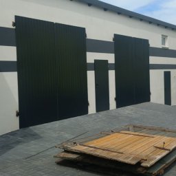 Kamil Jaskólski - Nowoczesny budynek z czarnymi, metalowymi bramami i drzwiami, ozdobiony poziomymi pasami. Na pierwszym planie sterta starych, drewnianych drzwi.