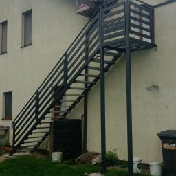 Kamil Jaskólski - Zewnętrzne, metalowe schody z platformą prowadzące do drzwi na piętrze budynku. Konstrukcja pomalowana na ciemny kolor, widok z dołu na tle zielonego trawnika.