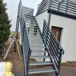 Kamil Jaskólski - Zewnętrzne schody metalowe z ażurowymi stopniami i szarą balustradą, prowadzące na balkon. Widoczne narzędzia i drabina sugerują świeży montaż.