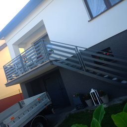 Kamil Jaskólski - Nowoczesna, pozioma balustrada metalowa w kolorze grafitowym, zamontowana na schodach zewnętrznych domu z szarą elewacją. Widoczne rośliny w donicach i fragment przyczepki samochodowej.