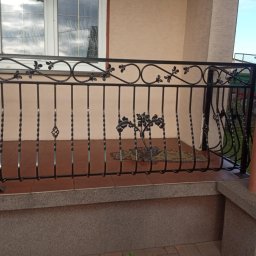 Kamil Jaskólski - Czarna, metalowa balustrada balkonowa z dekoracyjnymi elementami roślinnymi i spiralnymi prętami, zamontowana na brązowym balkonie z widocznym oknem w tle.