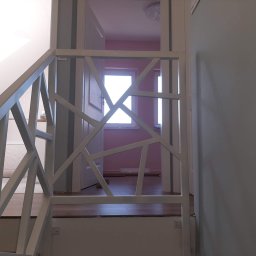 Kamil Jaskólski - Biała, metalowa balustrada schodowa z geometrycznym wzorem, widoczna z dołu schodów. W tle widoczne drzwi i fragment pokoju z oknem.