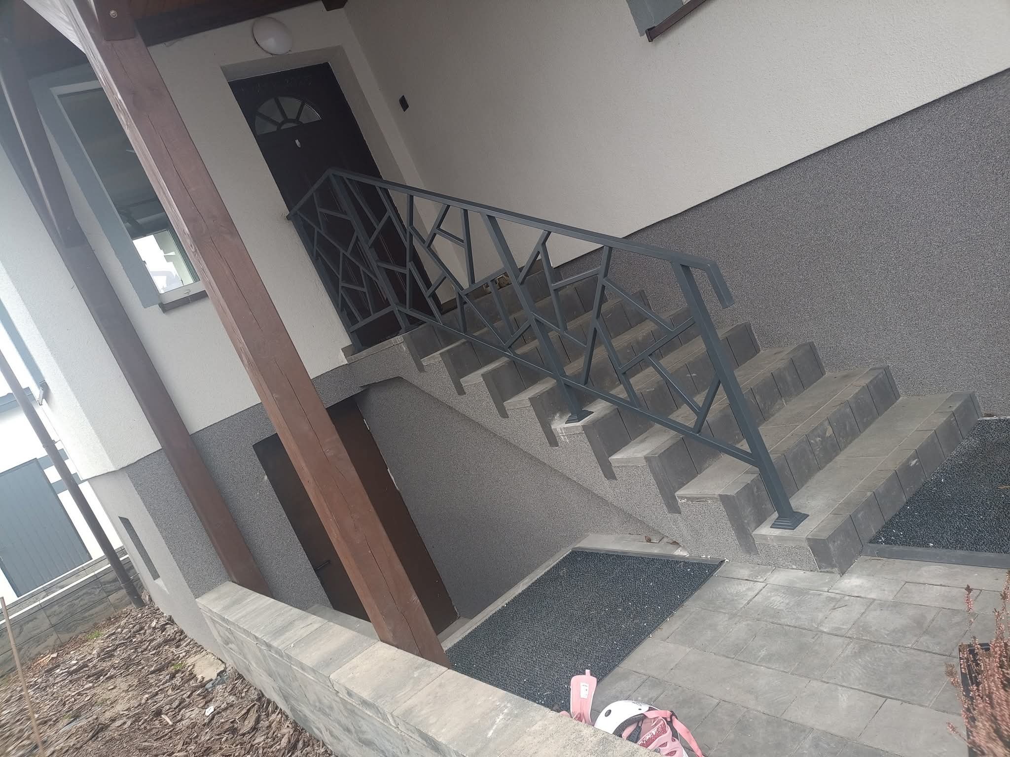 Schody zewnętrzne z geometryczną, metalową balustradą w odcieniach szarości, prowadzące do drzwi wejściowych. Widoczne elementy wykończenia elewacji i kostki brukowej.