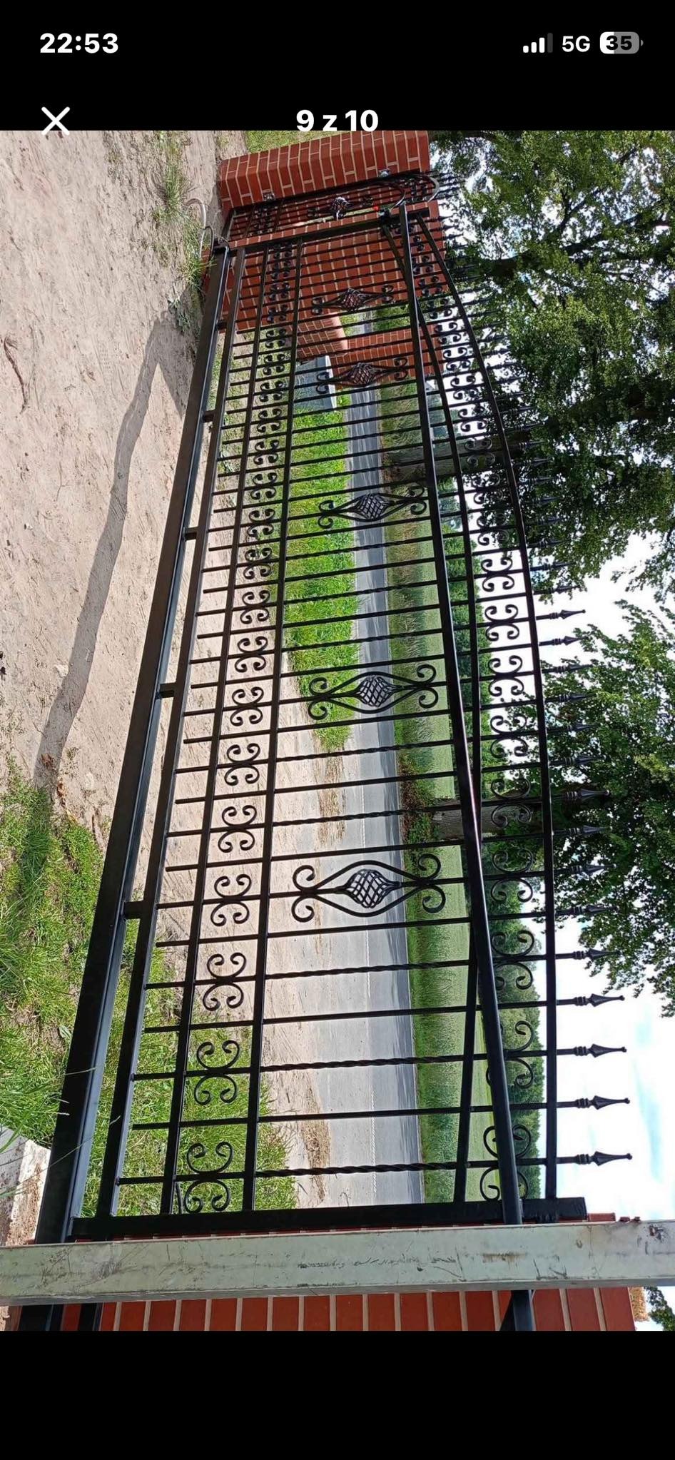 Czarna, metalowa brama z dekoracyjnymi elementami w kształcie wolut i rombów, zamontowana na ceglanym murku, widok z perspektywy ulicy.