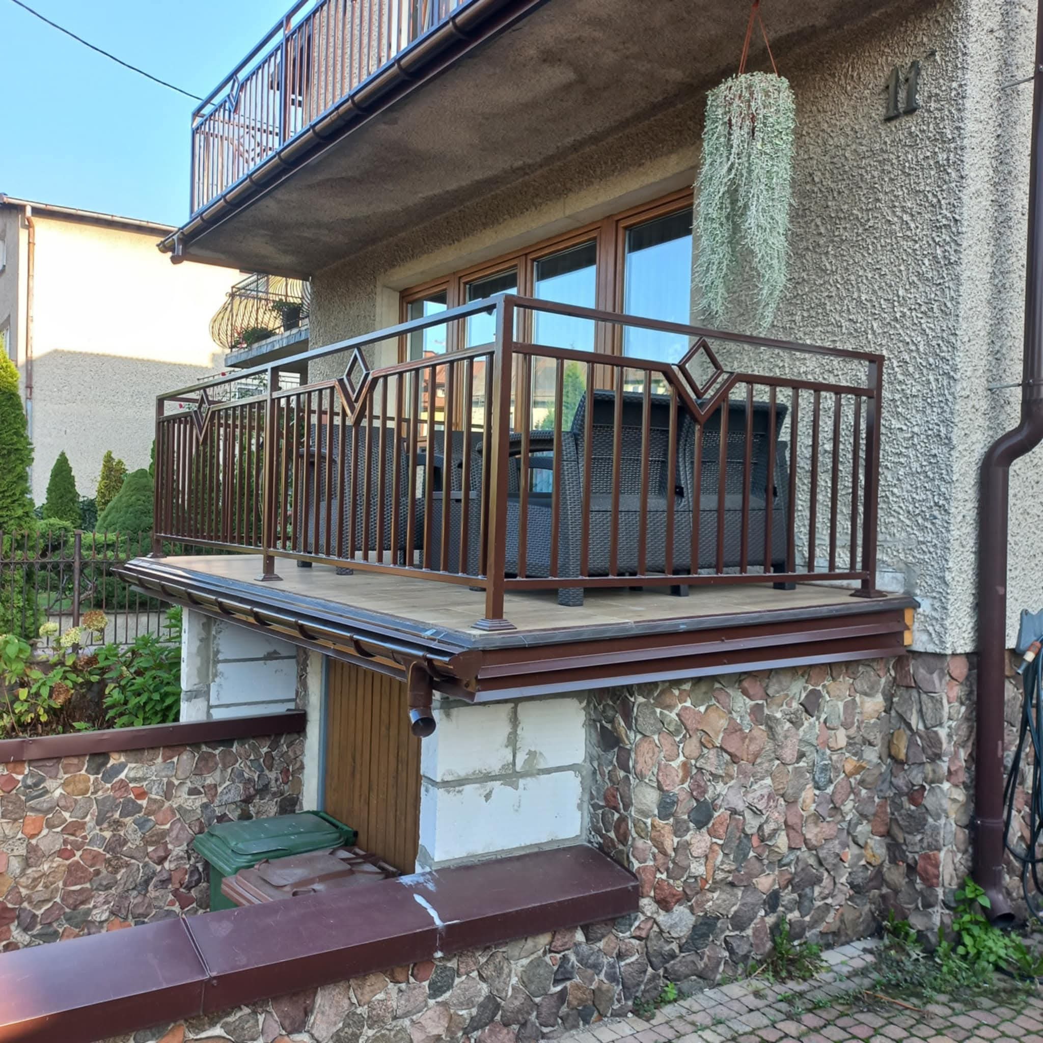 Balkon z brązową, metalową balustradą o geometrycznym wzorze rombów, z widocznymi meblami ogrodowymi. Kamienna podmurówka i wisząca roślina dodają uroku.