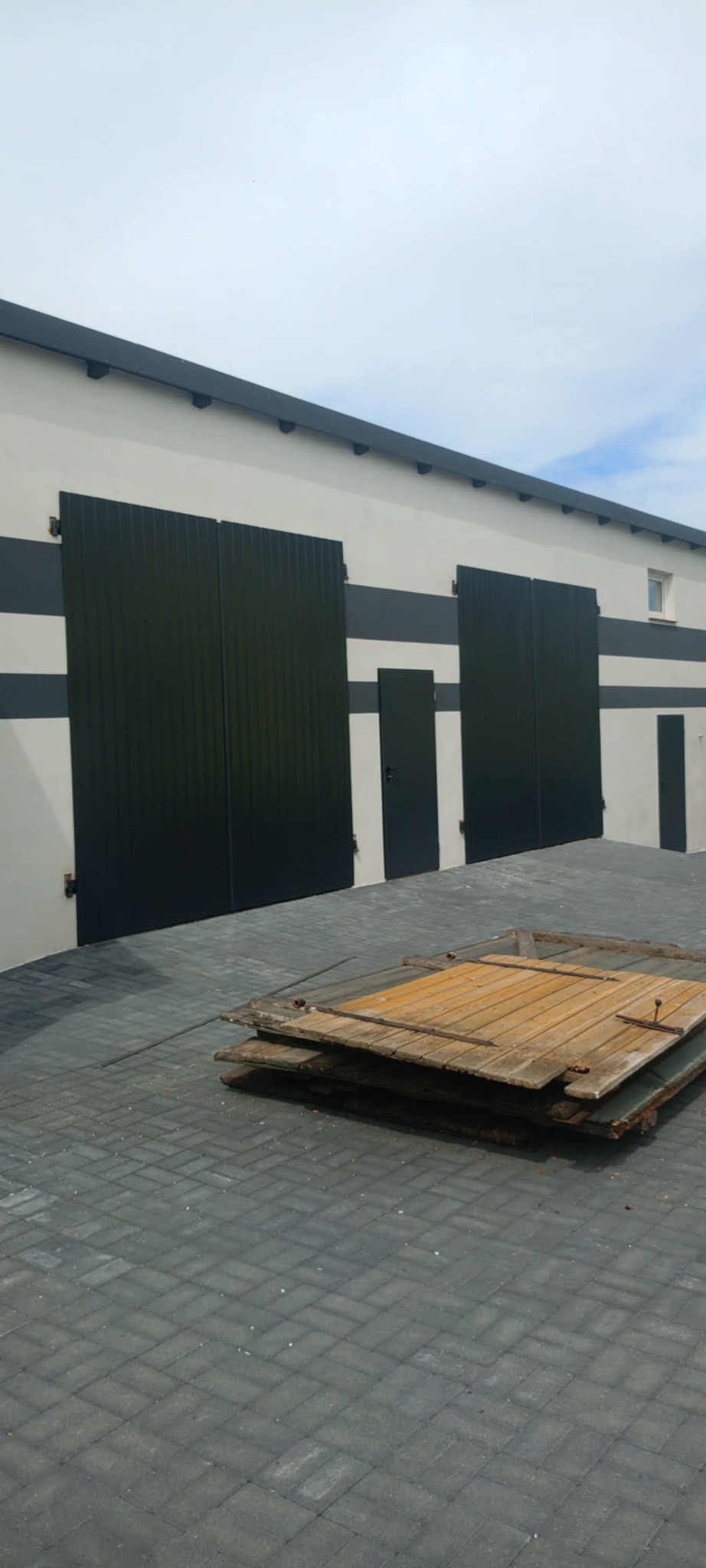 Nowoczesny budynek z czarnymi, metalowymi bramami i drzwiami, ozdobiony poziomymi pasami. Na pierwszym planie sterta starych, drewnianych drzwi.