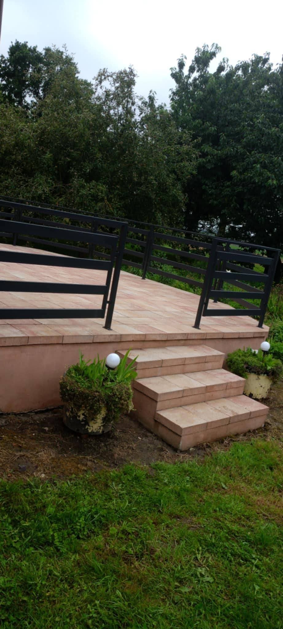 Taras z cegły klinkierowej z czarną, metalową balustradą. Schody prowadzą na podest, a w tle widoczne są drzewa i trawa. Oświetlenie w postaci kulistych lamp.