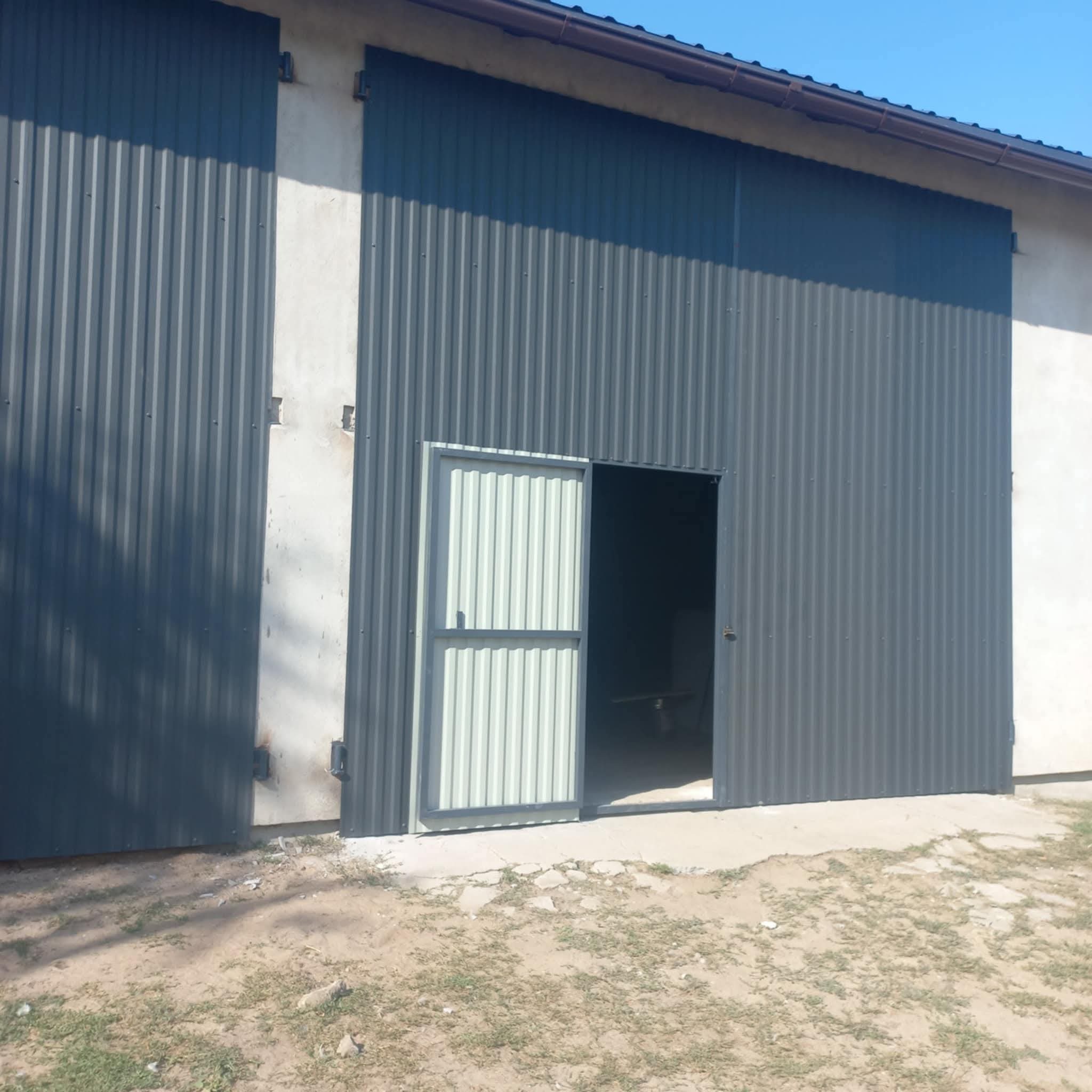 Szara, metalowa brama przesuwna z częściowo otwartymi drzwiami wejściowymi, wykonana z blachy falistej. Widoczne mocowania i fragment budynku.
