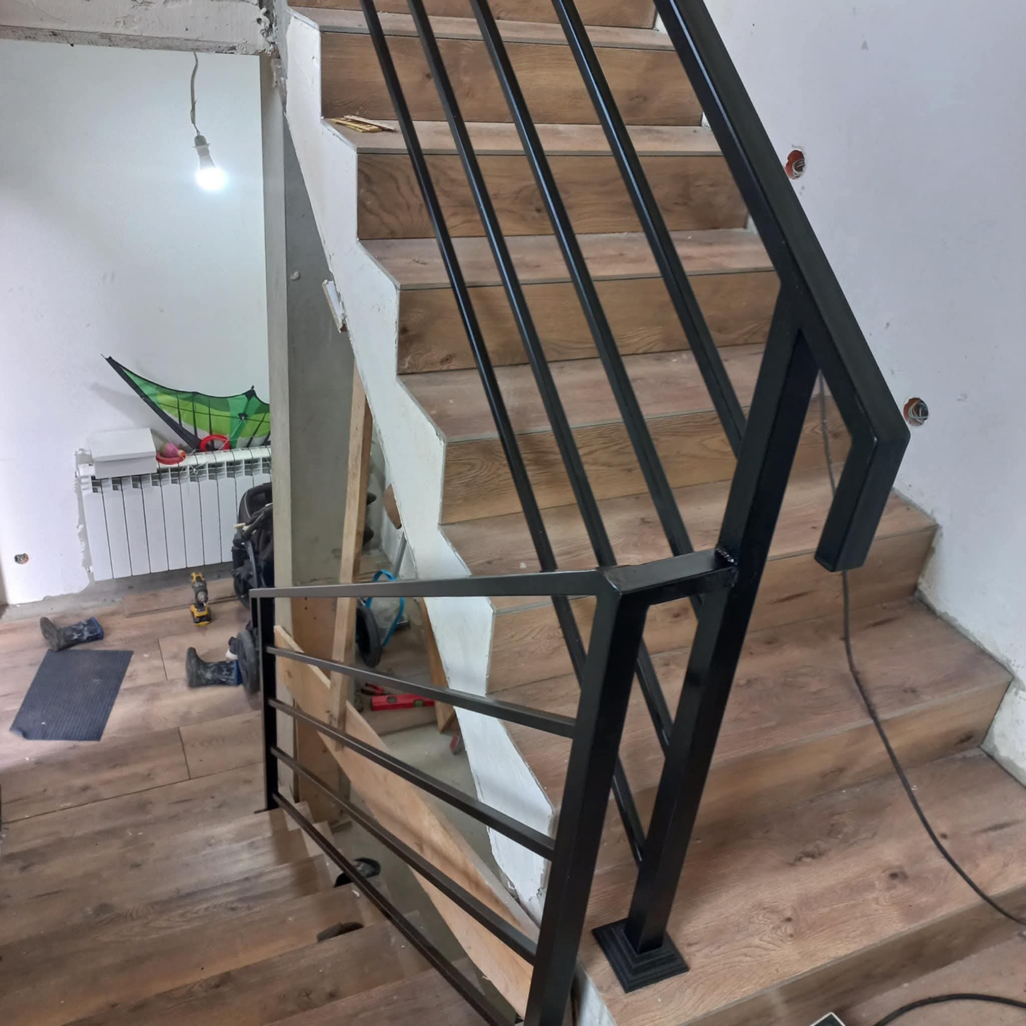 Nowoczesna, czarna balustrada metalowa z poziomymi prętami, zamontowana przy drewnianych schodach w surowym wnętrzu. Widoczne niedokończone wykończenia ścian.