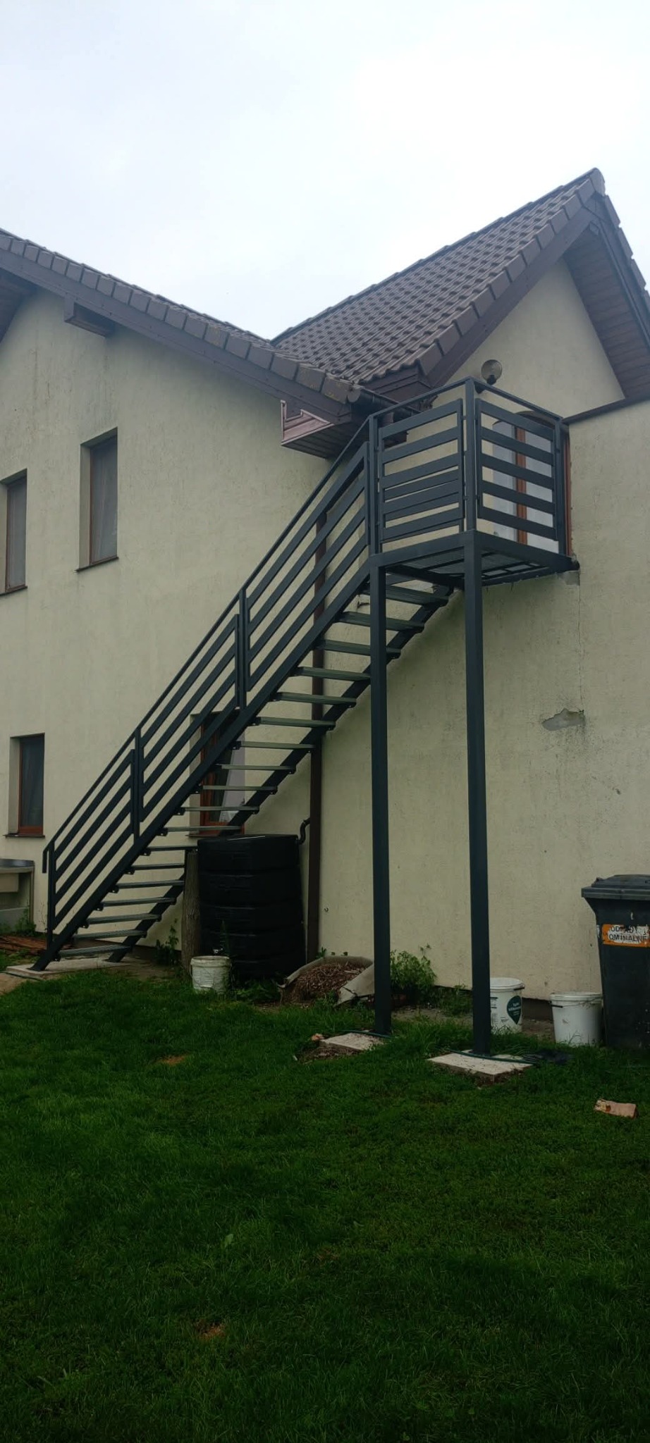 Zewnętrzne, metalowe schody z platformą prowadzące do drzwi na piętrze budynku. Konstrukcja pomalowana na ciemny kolor, widok z dołu na tle zielonego trawnika.