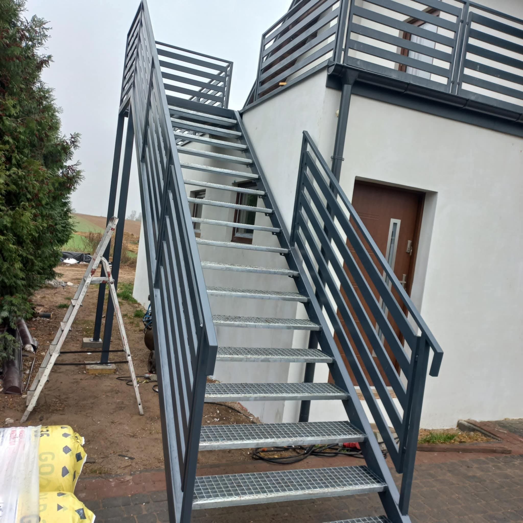 Zewnętrzne schody metalowe z ażurowymi stopniami i szarą balustradą, prowadzące na balkon. Widoczne narzędzia i drabina sugerują świeży montaż.
