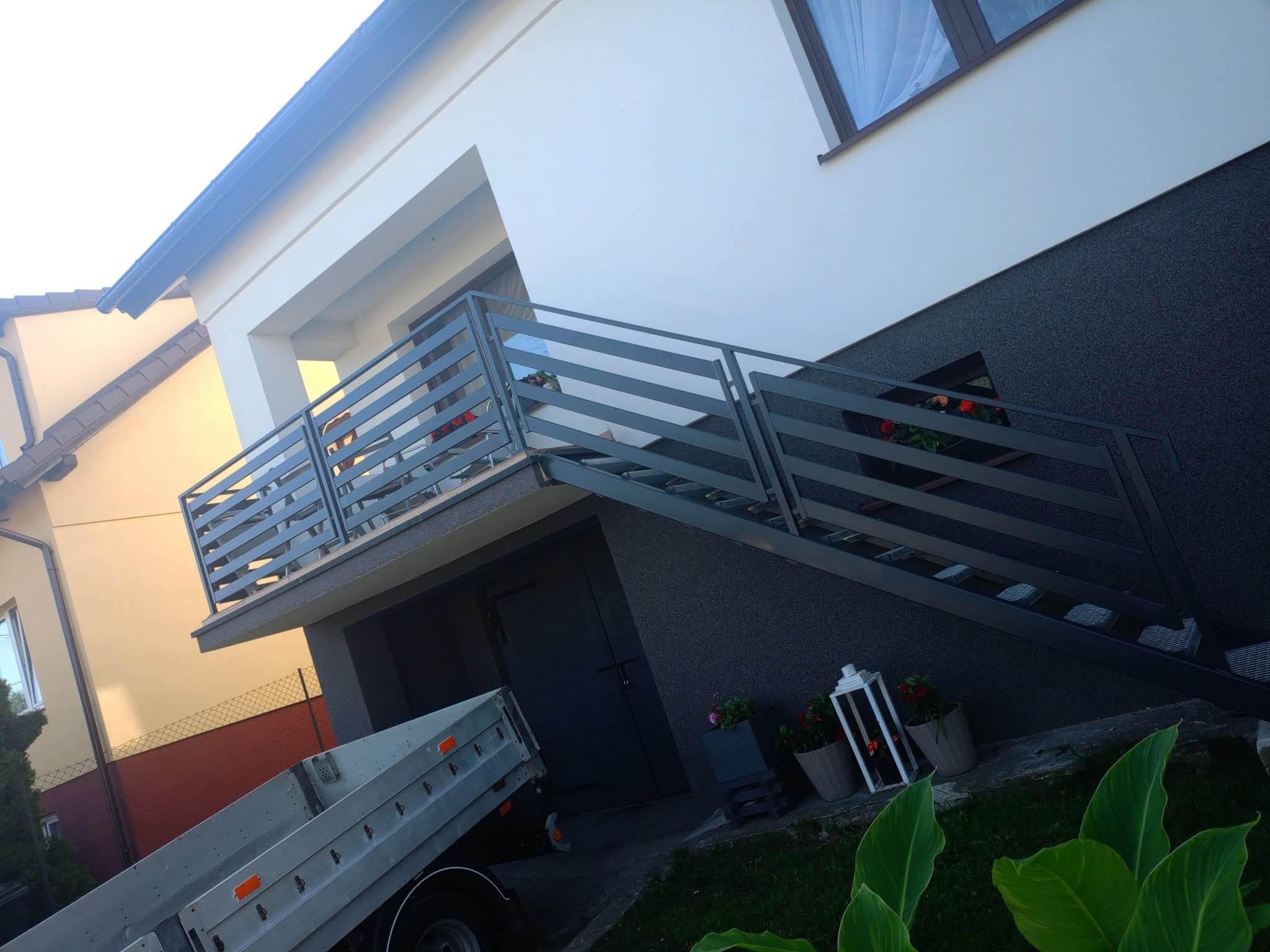 Nowoczesna, pozioma balustrada metalowa w kolorze grafitowym, zamontowana na schodach zewnętrznych domu z szarą elewacją. Widoczne rośliny w donicach i fragment przyczepki samochodowej.