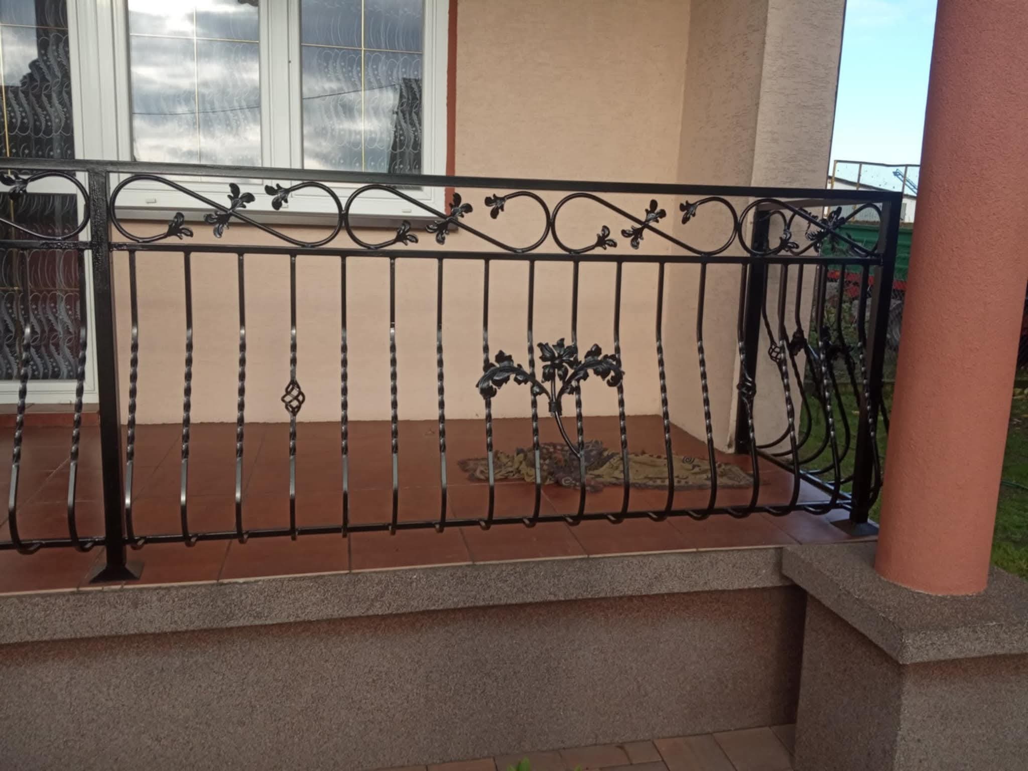 Czarna, metalowa balustrada balkonowa z dekoracyjnymi elementami roślinnymi i spiralnymi prętami, zamontowana na brązowym balkonie z widocznym oknem w tle.