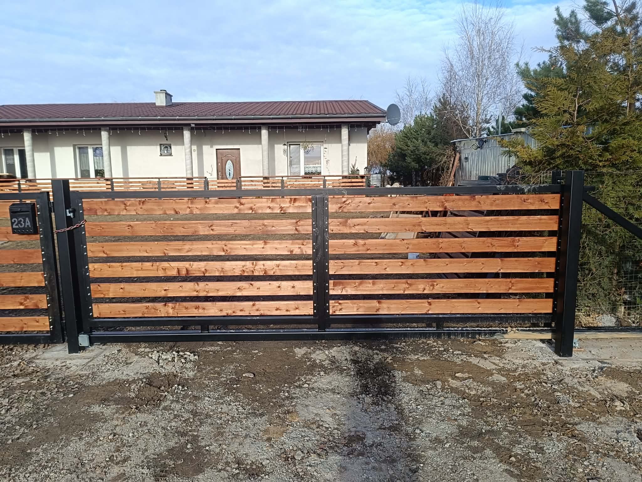 Dwuskrzydłowa brama wjazdowa z poziomych desek w ciepłym odcieniu drewna, osadzona w czarnej, metalowej ramie. Widoczny numer domu na skrzynce pocztowej.