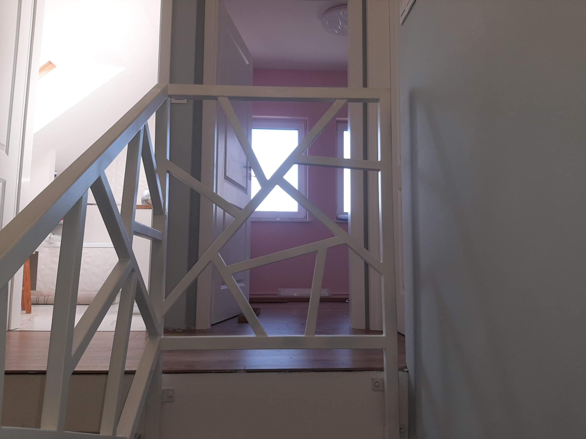 Biała, metalowa balustrada schodowa z geometrycznym wzorem, widoczna z dołu schodów. W tle widoczne drzwi i fragment pokoju z oknem.