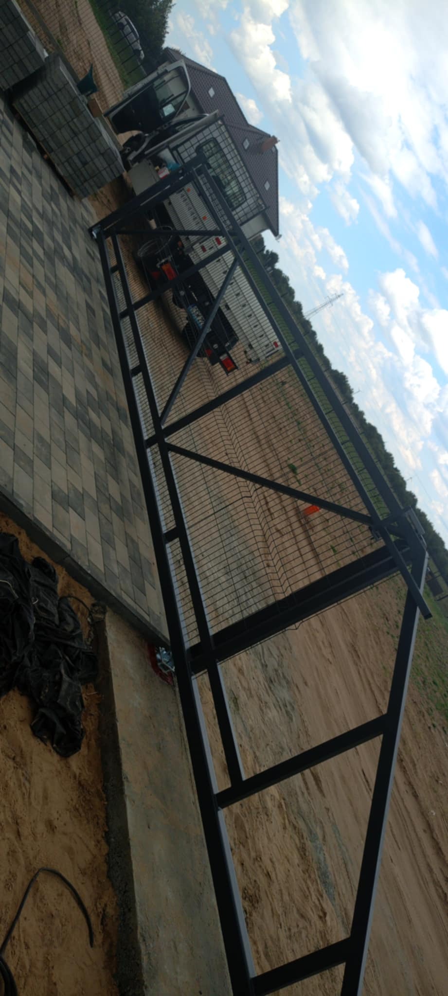 Częściowo zamontowane, czarne, metalowe ogrodzenie panelowe na tle piaszczystego terenu i błękitnego nieba. Widoczny samochód dostawczy i kostka brukowa.