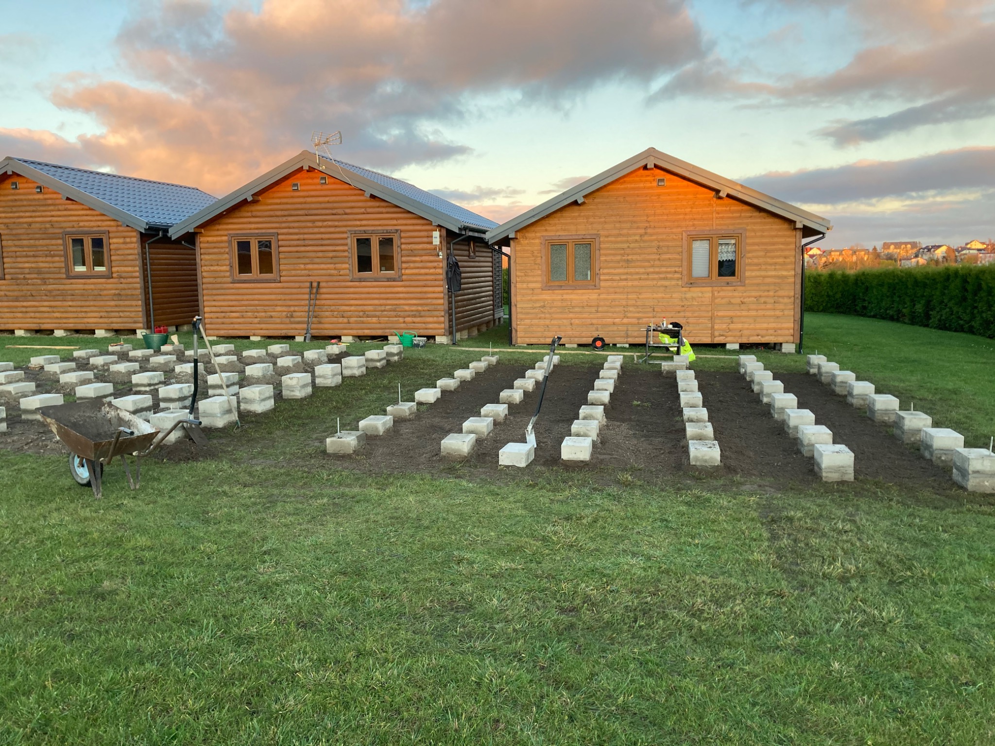 Fundamenty pod domki szkieletowe na trawie, wózek z ziemią i narzędzia. Widoczne trzy domki z drewna, niebo z różowymi chmurami o zachodzie słońca w tle.