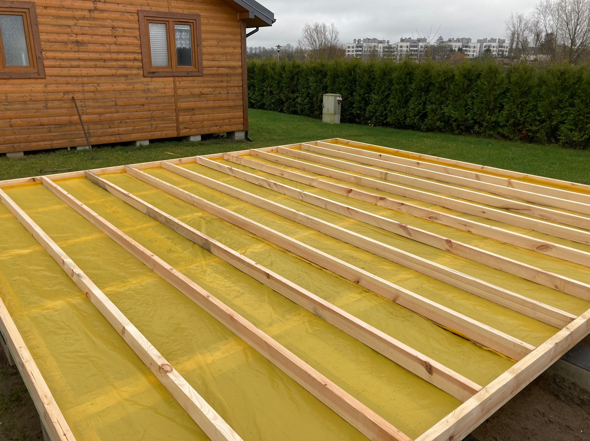 Konstrukcja podłogi z drewnianych belek na żółtej folii, widok z góry. W tle drewniany budynek i zieleń. Etap budowy domu szkieletowego.