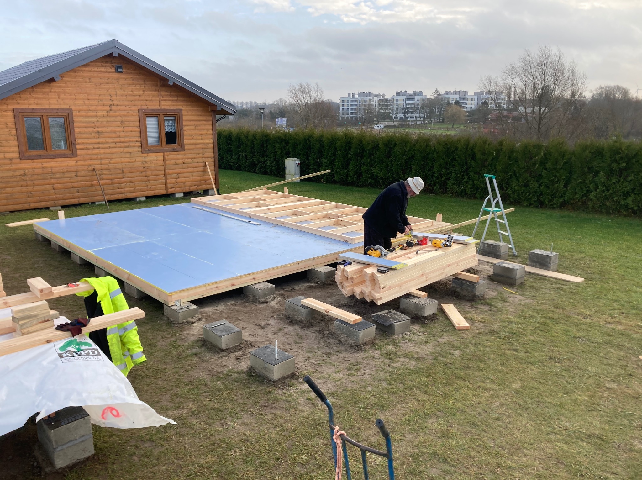 Budowa domu szkieletowego: montaż drewnianej konstrukcji podłogi na betonowych bloczkach. W tle gotowy drewniany domek i zabudowa miejska.