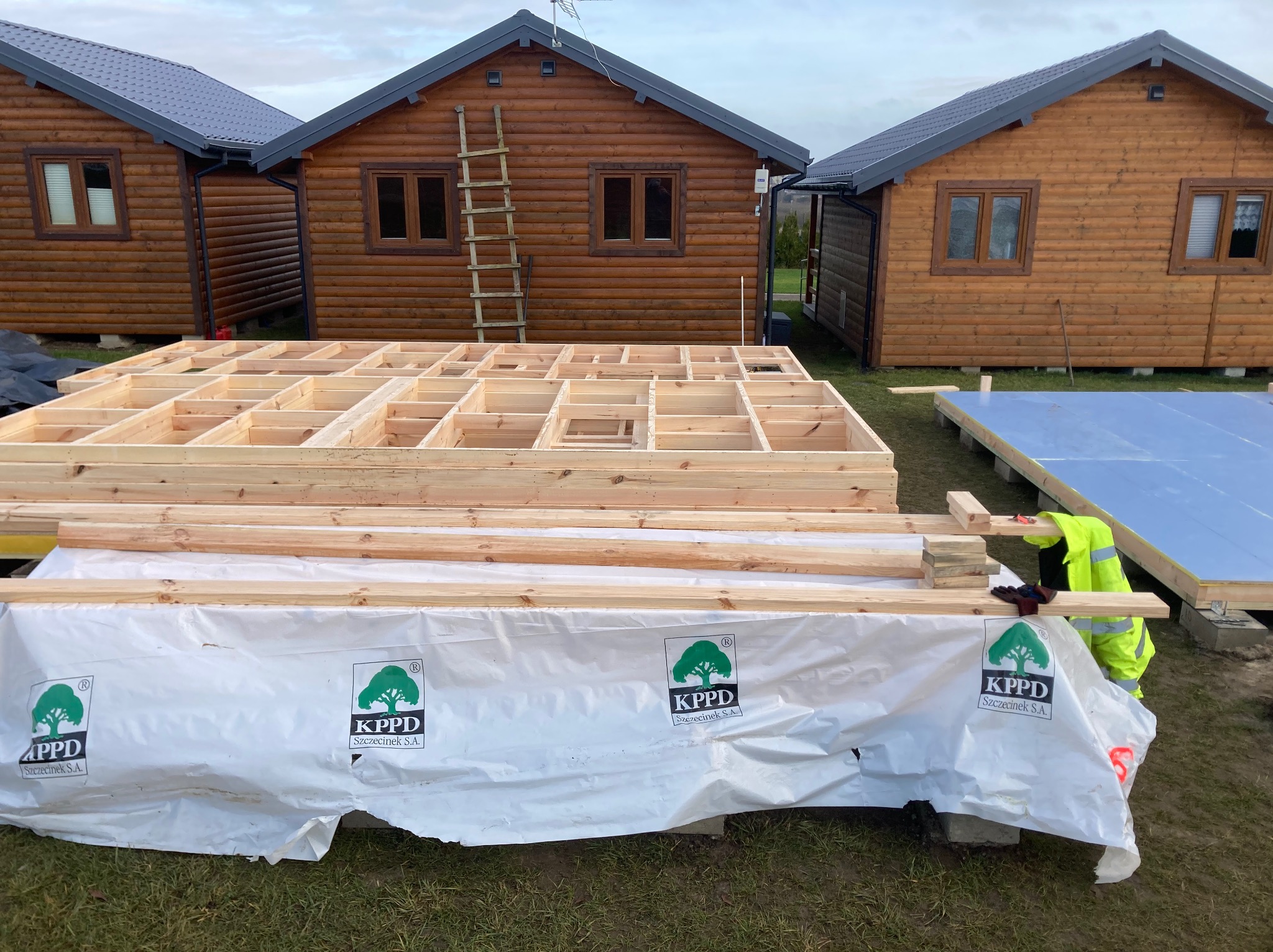 Konstrukcja szkieletowa domu z drewna na etapie budowy, widoczne elementy konstrukcyjne i materiały izolacyjne. W tle gotowe domki z drewna.