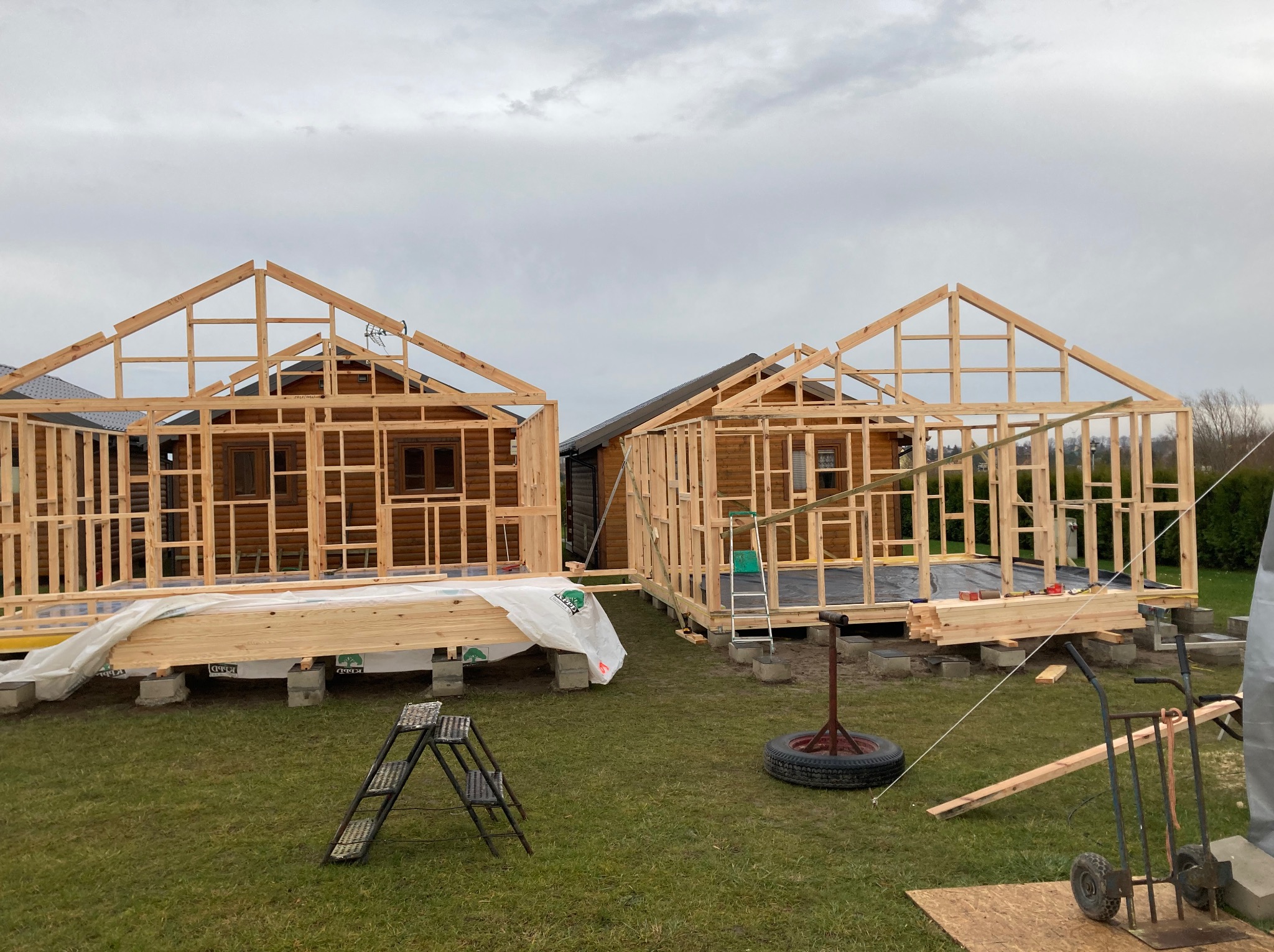 Dwa domy szkieletowe w trakcie budowy, widoczne drewniane ramy konstrukcyjne, narzędzia budowlane na trawie, pochmurne niebo w tle. Etap wznoszenia ścian i dachu.
