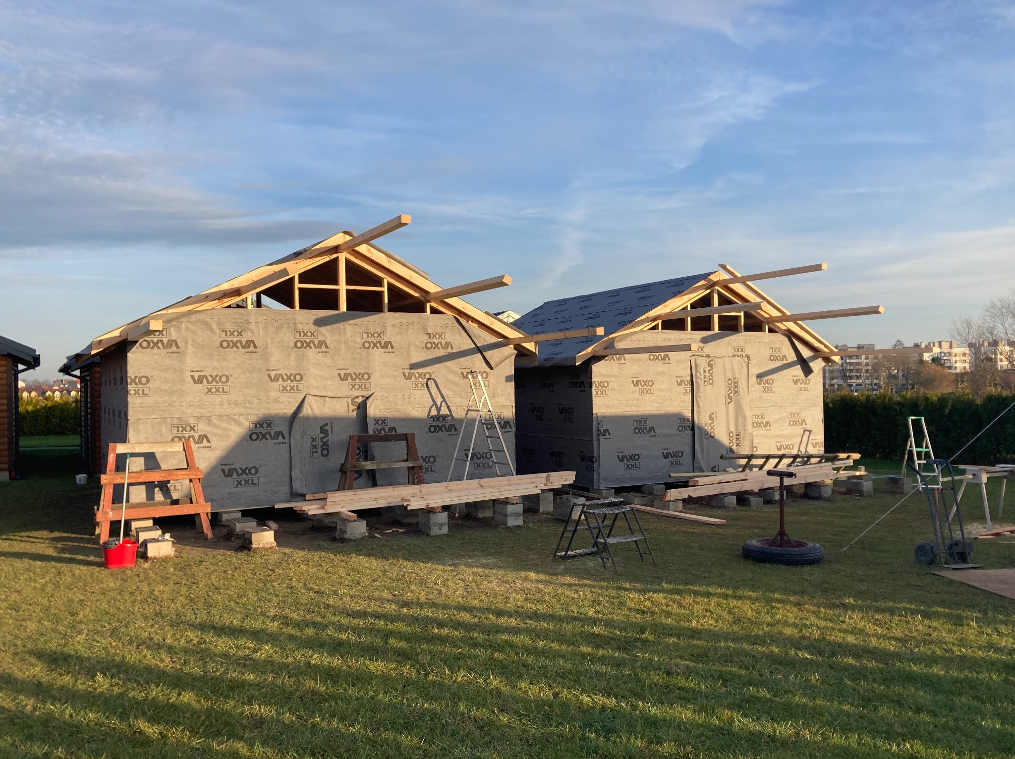 Dwa domy szkieletowe w trakcie budowy, zabezpieczone folią OXVA XXL, z widocznymi elementami drewnianej konstrukcji dachu i rusztowaniami na trawiastym terenie.
