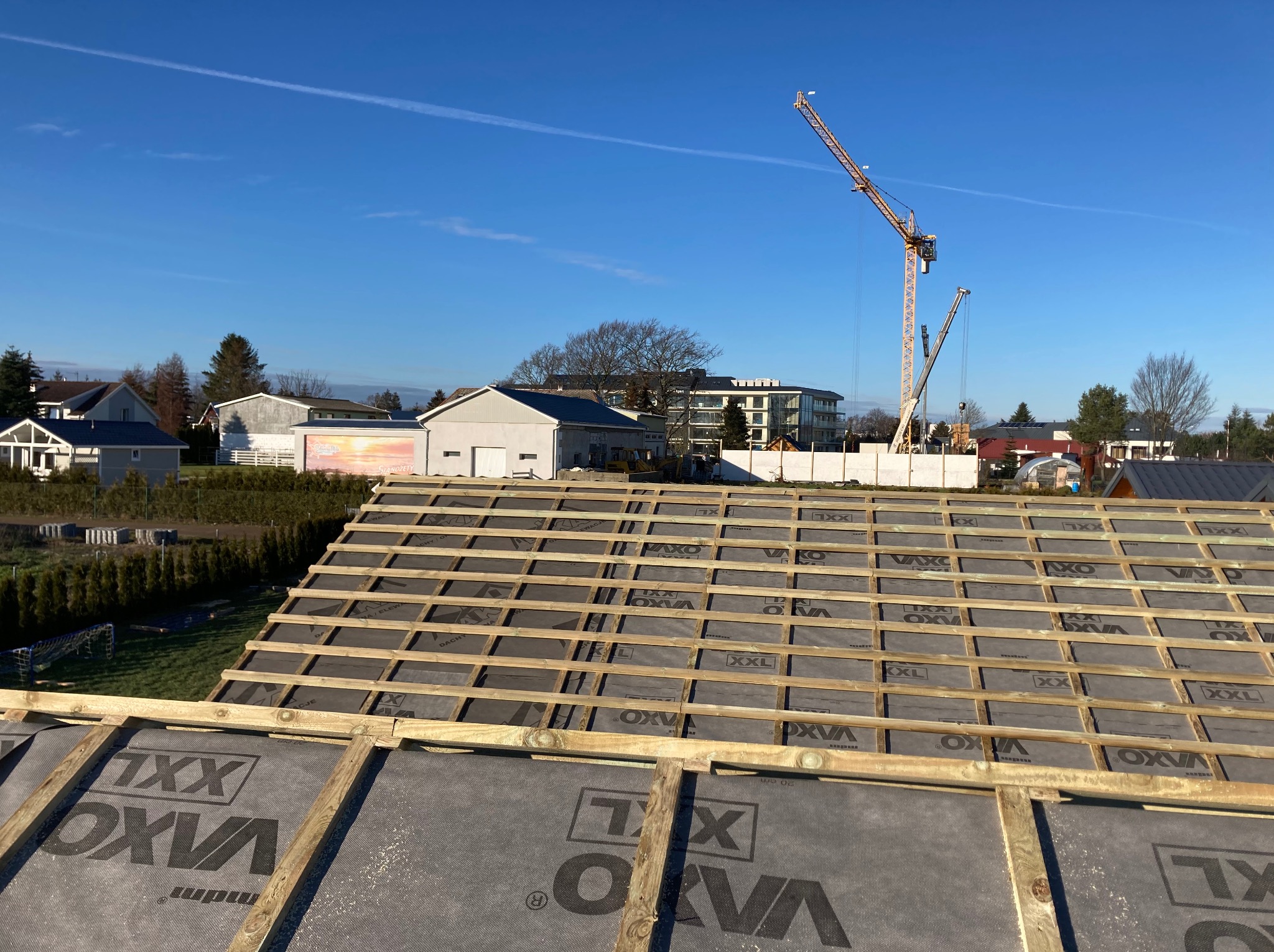 Konstrukcja dachu z łatami i membraną w trakcie budowy domu szkieletowego, widok z góry na tle zabudowy i żurawia budowlanego w słoneczny dzień.