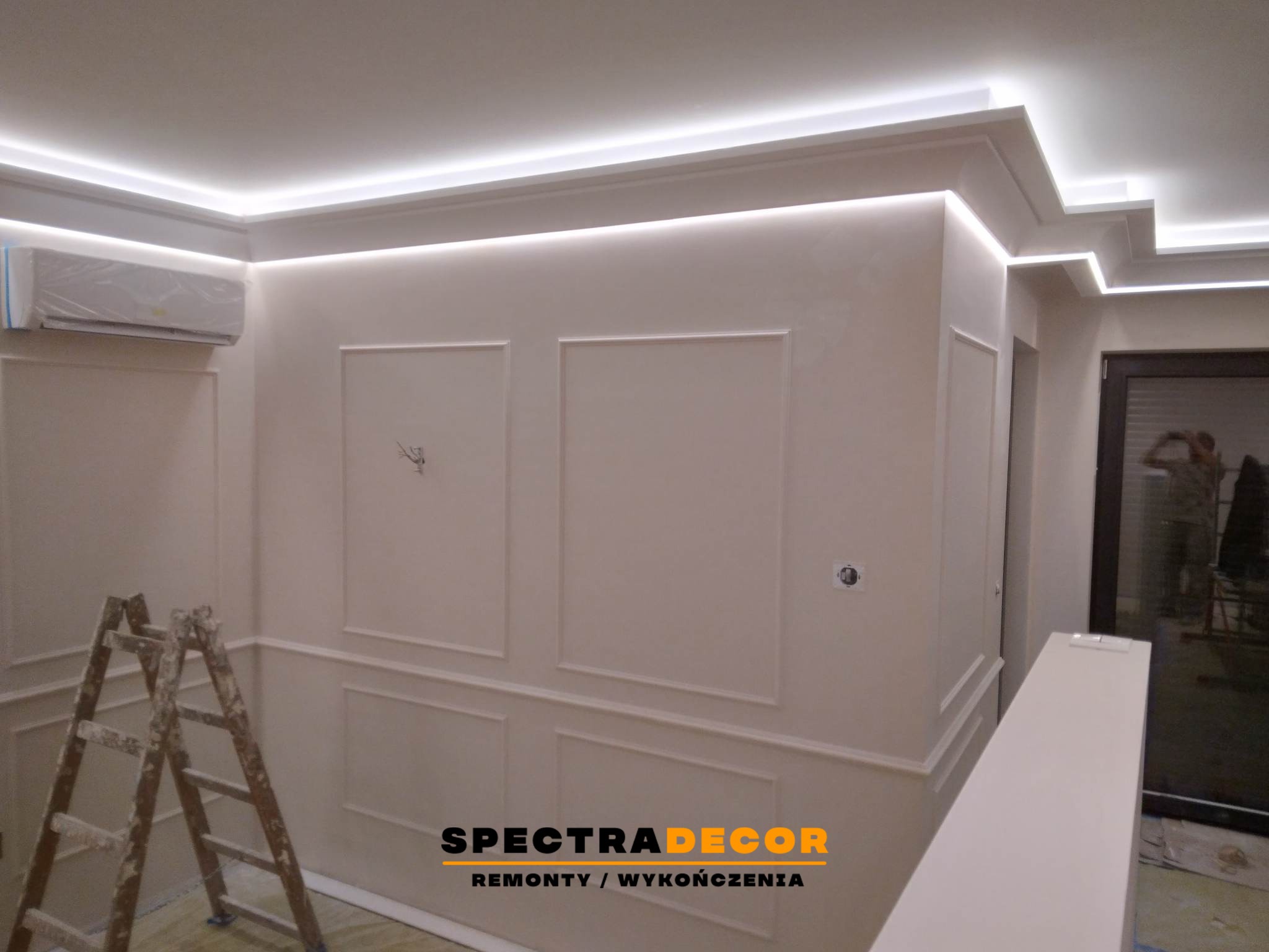 Wykończone wnętrze z dekoracyjnymi listwami ściennymi i oświetleniem LED podsufitowym. Widoczna drabina malarska i klimatyzator w folii ochronnej.
