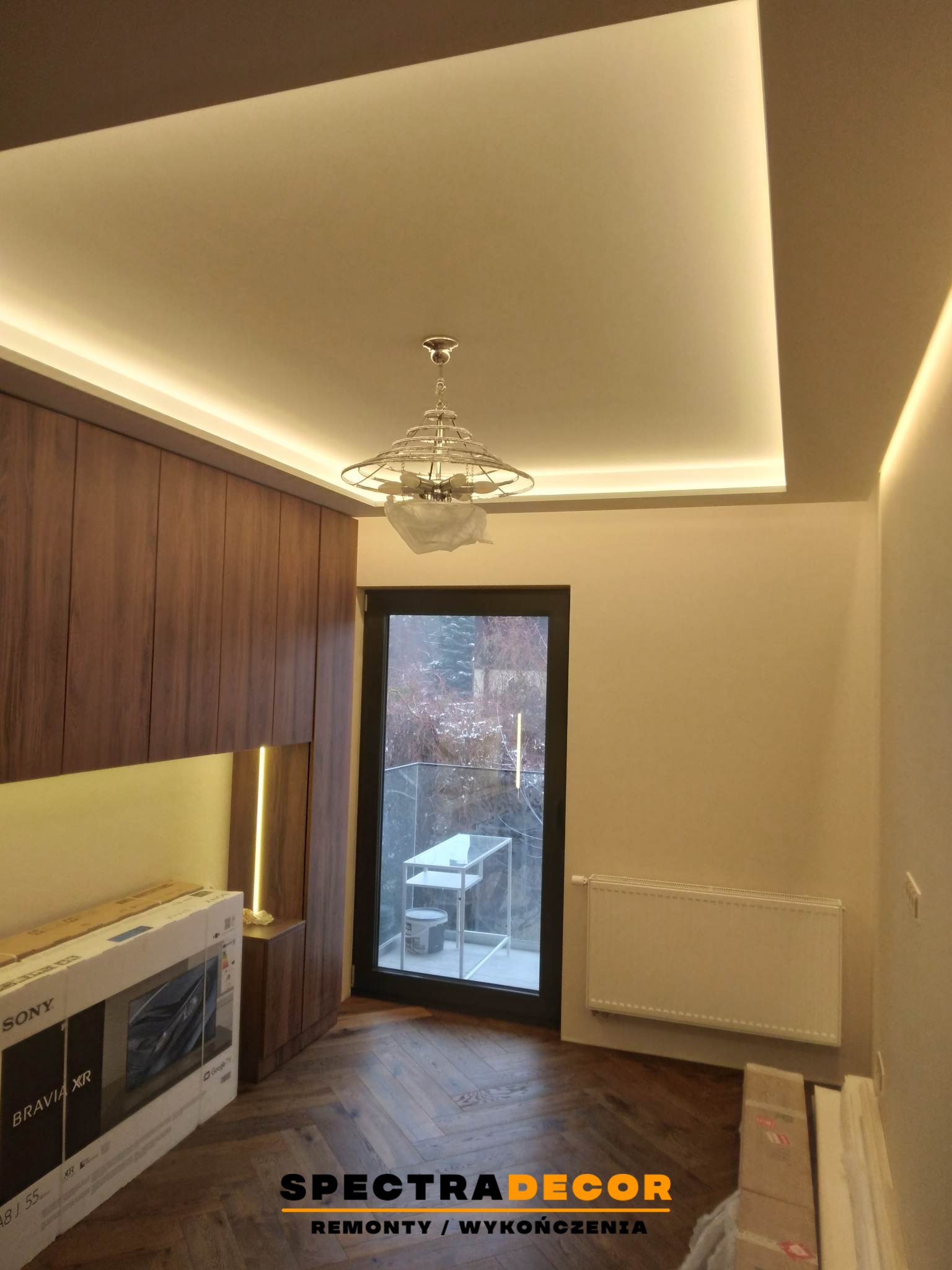 Nowoczesny salon po remoncie: drewniana zabudowa, parkiet w jodełkę, podwieszany sufit z oświetleniem LED i okno balkonowe z widokiem na zimowy krajobraz.